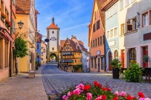 Bild der Altstadt von Rothenburg ob der Tauber in Bayern, Deutschland. Kopfsteinpflasterstraße, umgeben von bunt bemalten Häusern mit Fachwerk und Blumen im Vordergrund. Im Hintergrund ein historischer Turm.