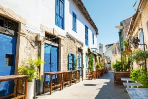 Gasse mit historischen Gebäuden, blauen Fensterläden und Außenterrasse bei Sonnenschein.