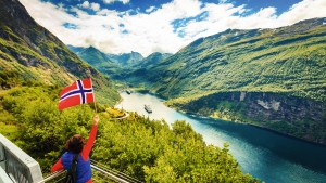 Person schwenkt norwegische Flagge vor Fjordlandschaft.