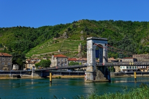 Hängebrücke über Fluss vor hügeliger Landschaft mit Weinbergen und Gebäuden.