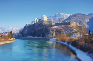 Burg auf einem Hügel an einem Fluss im winterlichen Alpenpanorama.