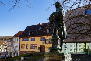 Bronzestatue eines Mannes vor historischem Gebäude und modernen Glasbau im Hintergrund.