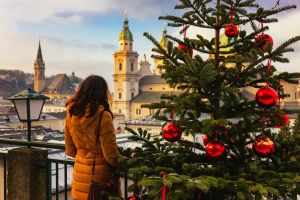 Person in Winterjacke steht neben geschmücktem Weihnachtsbaum, Blick auf Stadt im Hintergrund.
