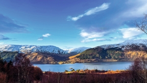 Berglandschaft mit See, bewaldeten Hängen und teilweise schneebedeckten Gipfeln.