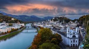 Panoramablick auf eine Stadt mit Fluss, Brücke, historischen Gebäuden und bewölktem Himmel.