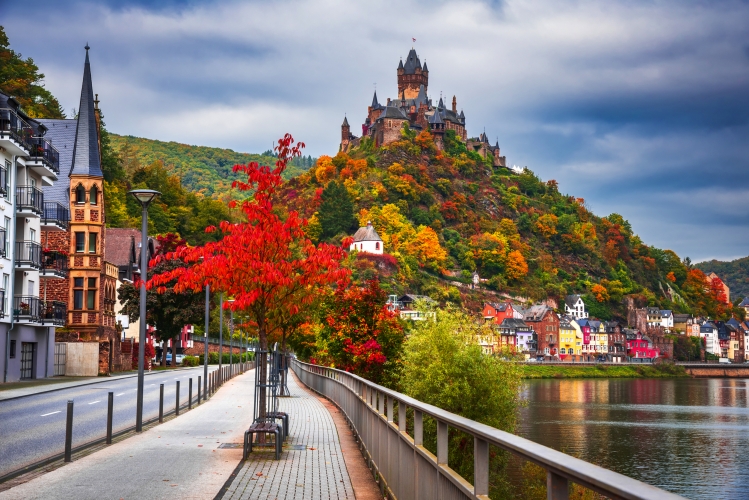 Burg auf Hügel mit Herbstlaub, Fluss und bunten Häusern im Vordergrund.