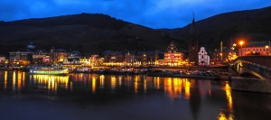 Beleuchtete Stadtansicht am Flussufer bei Nacht mit Hügeln im Hintergrund.