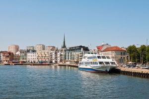 Ein Fluss mit Anlegestelle, an der ein großes Passagierschiff liegt. Im Hintergrund ist eine städtische Skyline mit mehreren mehrstöckigen Gebäuden und einem spitzen Kirchturm zu sehen. Der Himmel ist klar und blau.