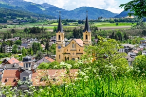 Kirche mit zwei Türmen in Alpenstadt, umgeben von Hügeln und Berglandschaft.