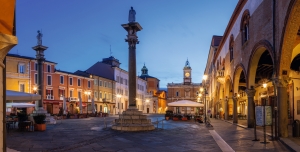 Städtischer Platz mit Säule und Gebäuden bei Abenddämmerung.