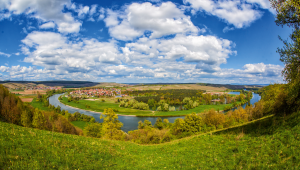 Landschaft mit Flussbiegung, grünen Wiesen, Bäumen und blauem Himmel mit Wolken.