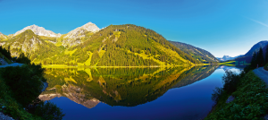 Eine Berglandschaft mit einem See im Vordergrund, der die umliegenden Berge und den klaren blauen Himmel widerspiegelt. Die Berge sind teilweise von grünen Wäldern bedeckt, und die Szene vermittelt eine ruhige und friedliche Atmosphäre.