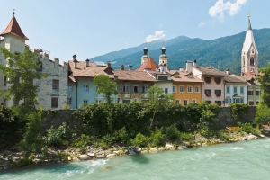 Bunte Häuserreihe am Fluss mit Bergkulisse im Hintergrund und einem Kirchturm.