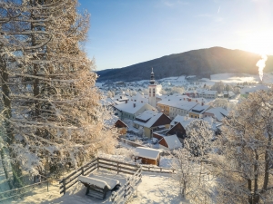 Winterlandschaft mit verschneitem Dorf und Kirchturm im Sonnenlicht.