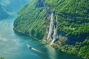 Ein Wasserfall fließt eine grüne Felswand hinab, ein Boot fährt auf einem Fjord vorbei.