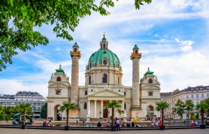 Barockkirche mit grüner Kuppel und zwei Säulen, umgeben von Bäumen und Leuten.