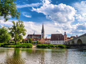 Stadtansicht mit Fluss, Kirche und Steinbrücke im Vordergrund bei blauem Himmel.