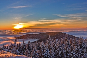 Winterlandschaft mit Sonnenuntergang über schneebedecktem Wald und Wolkenmeer.