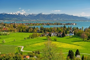Blick auf Landschaft mit Bodensee, grünen Feldern und schneebedeckten Bergen im Hintergrund.