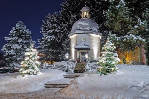Verschneite Kapelle bei Nacht mit beleuchteten Weihnachtsbäumen.