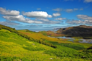 Grüne Hügel mit üppiger Vegetation erstrecken sich bis zu einem ruhigen Meereshorizont unter einem klaren blauen Himmel mit weißen Wolken. Im Hintergrund sind felsige Erhebungen zu sehen, die dem Landschaftsbild Tiefe verleihen.