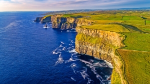 Luftaufnahme der Cliffs of Moher in Irland, die aus der blauen, sich schlängelnden Küste aufragen und von grünen Feldern umgeben sind.