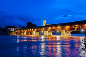 Beleuchtete Holzbrücke über einen Fluss bei Nacht, reflektiert im Wasser.