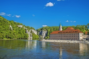 Kloster an einem Flussufer mit bewaldeten Hügeln im Hintergrund.