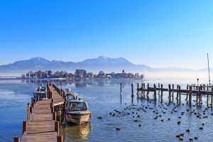Hölzerner Steg mit Booten auf einem See, Berge im Hintergrund, viele Wasservögel.