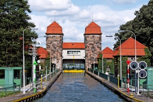 Schleuse mit zwei steinernen Gebäuden und roten Dächern, umgeben von Bäumen unter blauem Himmel.